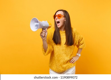 Portrait of funny pretty young woman in fur sweater, orange heart eyeglasses screaming on megaphone isolated on bright yellow background. People sincere emotions, lifestyle concept. Advertising area