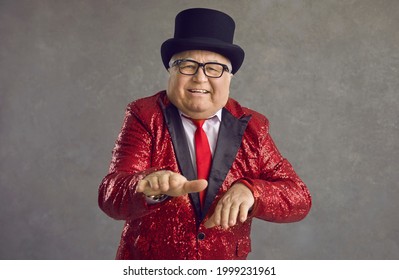 Portrait of funny rich chubby senior man dancing on gray studio background. Happy fat millionaire in red retro style sequin outfit and black tophat enjoying music and having fun at crazy disco party