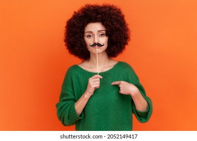 Portrait of funny serious woman with Afro hairstyle wearing green casual style sweater pointing at herself and holding paper mustache. Indoor studio shot isolated on orange background.