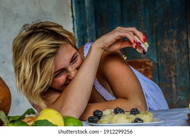 Portrait Of A Funny Sexy Blonde Woman With Blond Hair Eating At A Table On The Porch Of An Old Village House. A Bouquet Of Sunflowers.