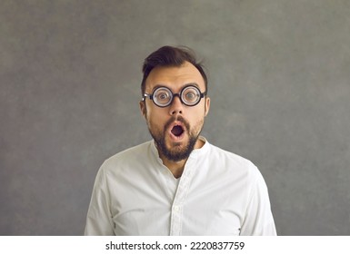 Portrait Of Funny Shocked Man Round Hilarious Glasses Standing On Grey Background. Puzzled Bearded Adult Businessman Staring Eyes And Opened Mouth Looking At Camera. Human Face Expression And Reaction