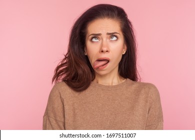 Portrait Of Funny Silly Young Woman With Brunette Hair Looking Cross Eyed And Sticking Tongue Out, Making Dumb Brainless Face, Disobedient Goofy Expression. Studio Shot Isolated On Pink Background