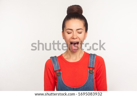 Portrait of funny stylish pretty girl with hair bun in denim overalls demonstrating tongue out expressing disobedience, derisive naughty expression. indoor studio shot isolated on white background