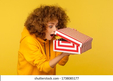 Portrait Of Funny, Surprised, Curious Curly-haired Woman In Hoodie Looking Into Gift Box, Unwrapping Present And Peeking Inside With Interest, Open Mouth In Amazement. Indoor Studio Shot Isolated