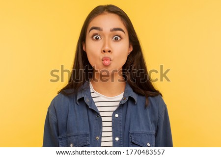 Portrait of funny surprised girl in denim shirt making fish face with pout lips and looking amazed, choking with idiotic comical silly expression. indoor studio shot isolated on yellow background