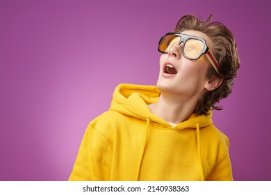 Portrait of a funny surprised teen boy in bright yellow hoodie looking up with surprise and excitement. Bright purple background with copy space. Education. Youth lifestyle, success. Emotions.