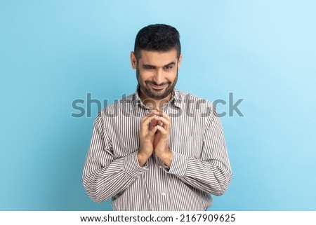 Portrait of funny tricky handsome businessman thinking devious plan with cunning face expression, looking at camera, wearing striped shirt. Indoor studio shot isolated on blue background.