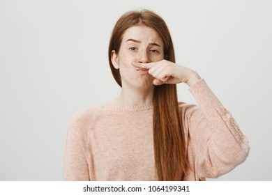 Portrait Of Funny Upbeat Ginger Girl Holding Index Finger Above Nose As If It Is Moustache And Lifting One Eyebrow, Copying Someone, Standing Over Gray Background. Student Makes Fun Of Her Teacher