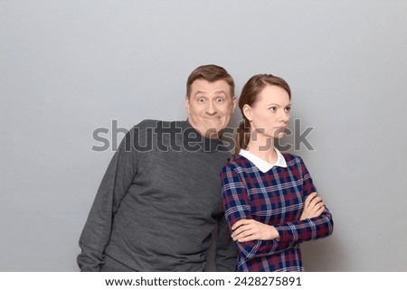 Portrait of funny weird couple, man is making goofy ridiculous faces, having fun, fooling around, woman is turned away from man, she is offended and serious, over gray background. Relationship concept