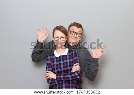 Portrait of funny weird couple, woman with serious expression, man is making goofy ridiculous faces, having fun, fooling around behind her, both are standing over gray background. Relationship concept
