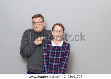 Portrait of funny weird couple, woman is making goofy ridiculous grimaces, man is pointing at her with finger, both are having fun, fooling around, standing over gray background. Relationship concept