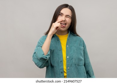 Portrait Of Funny Woman Putting Finger Into Her Nose And Showing Tongue, Fooling Around, Bad Habits, Disrespectful Behavior, Wearing Casual Style Jacket. Indoor Studio Shot Isolated On Gray Background