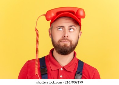 Portrait of funny worker man holding handset on his head and looking up, tired of calling for order service, wearing overalls and red cap. Indoor studio shot isolated on yellow background.