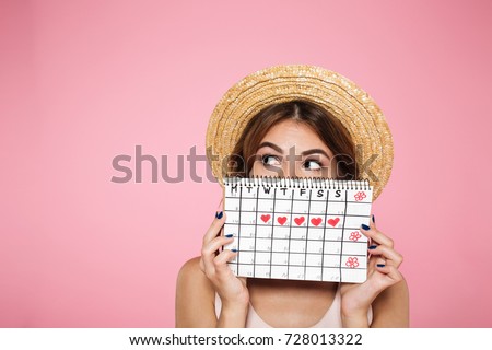 Portrait of a funny young girl in summer hat hiding behind a periods calendar and looking away at copy space isolated over pink background