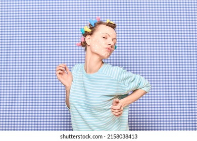 Portrait of funny young woman with colorful hair curlers on head, with skincare product on face, having fun, fooling around, pouting lips, grimacing ridiculously, over shower curtain background - Shutterstock ID 2158038413