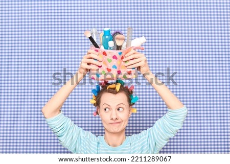Portrait of funny young woman with colorful hair curlers on head, holding makeup bag with beauty treatment accessories and cosmetics, making goofy face, fooling arounds over shower curtain background