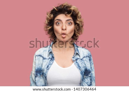 Portrait of funny young woman with curly hairstyle in casual blue shirt standing with fish pout lips and looking at camera with big eyes. indoor studio shot, isolated on pink background.