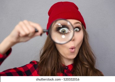 Portrait of a funny young woman looking at the camera through magnifying glass over gray background