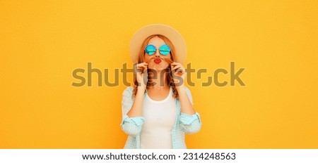 Portrait of funny young woman showing mustache her hair blowing lips with red lipstick sending air kiss wearing straw hat on orange background