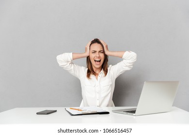 Portrait Of A Furious Young Business Woman Sitting At The Office Desk And Yelling Isolated Over White Background