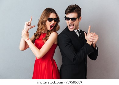 Portrait of an furious young couple dressed in formal wear and sunglasses standing back to back and showing gun gesture over gray wall background