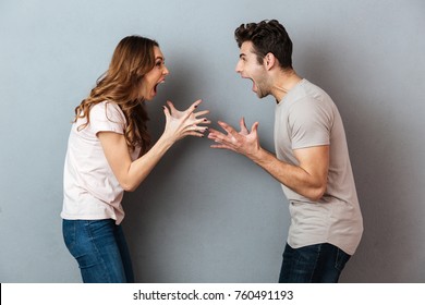 Portrait of a furious young couple having an argument while gesturing and looking at each other over gray wall