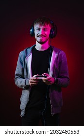 Portrait Of Gamer Playing Using A Console. Gamer Wearing Headset Standing And Holding White Console On Dark With Red Neon Lights Background