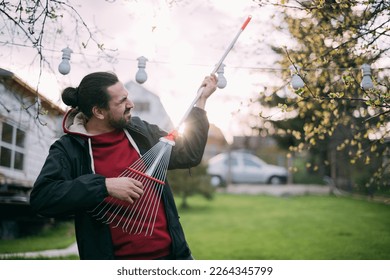 Portrait of a gardener with a fan rake in his hands, as if with a guitar in the garden at sunset. A man in work clothes holds a rake in his hands and pretends to play it like an electric guitar