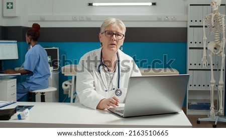 Portrait of general practitioner using laptop at health care checkup visit, working on appointment paperwork. Female physician looking at diagnosis results on medical system online.