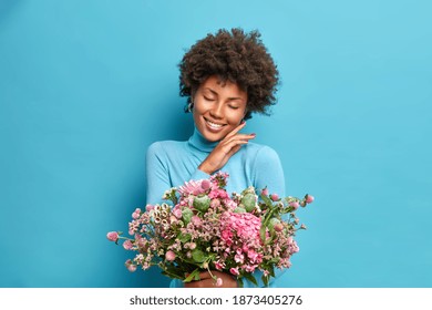 Portrait Of Gentle Dark Skinned Woman Touches Jawline Gently Closes Eyes And Smiles Pleasantly Holds Beeautiful Bouquet Of Flowers Has Romantic Mood Isolated Over Blue Background. Feminine Concept