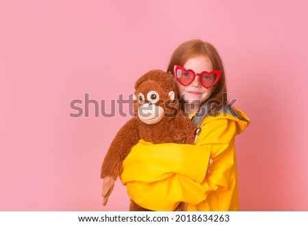 Portrait of ginger kid girl playing with stuffed animal pet. Funny child with toy monkey isolated on pink background. Orange toy and kid.