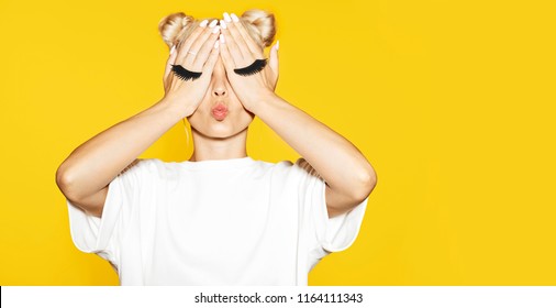 Portrait of girl with blond hair sending air kiss with fake eyelash on hands on yellow background. Concept of emotions. 