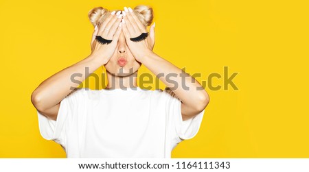Portrait of girl with blond hair sending air kiss with fake eyelash on hands on yellow background. Concept of emotions. 