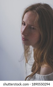 Portrait of a girl with blonde long hair and freckles on her face without makeup. A young woman dressed in a white sweater sitting against a white wall and has sad looking smile.