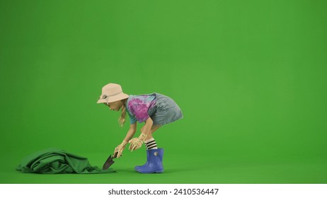 Portrait of girl in dress and rubber boots on chroma key green screen. Small cute girl gardener planting seeds in soil with shovel.