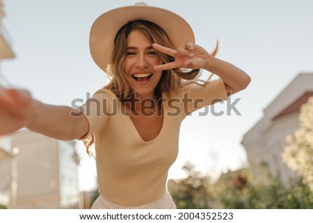 Portrait of girl of European appearance shows sign of peace and smiles broadly against background of sky. She is wearing light beige blouse and stylish hat.