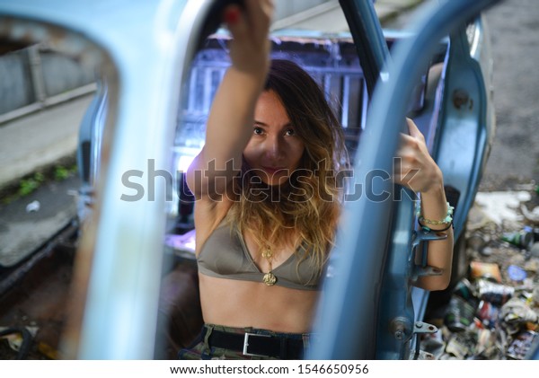 portrait of girl in green bra,\
camouflage print pants and necklace near the blue old car in the\
street