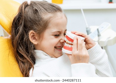 Portrait of a girl holding a model of a jaw with teeth while sitting in a dental office, playing and grimacing. Children's dentistry. Children's teeth care. - Powered by Shutterstock