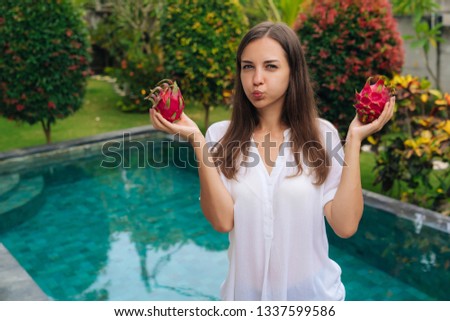 Portrait girl holds two dragon fruits, pitaya in her hands and makes funny face inflates her cheeks. Cute girl standing with fresh fruits background of pool. Concept healthy food, vitamins, diet