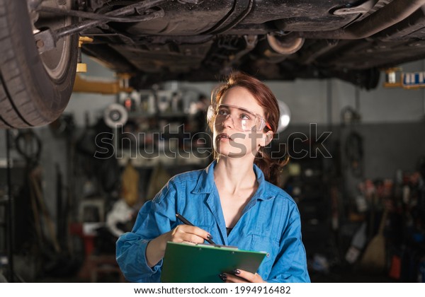 Portrait of a girl in overalls and glasses in a car\
repair shop.Blue overalls on a woman car mechanic. The concept of\
diagnostics and repair of machines.A specialist in a garage with a\
car makes notes