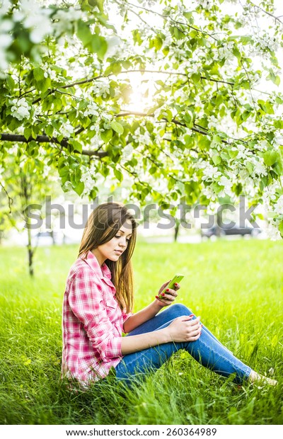 Portrait of girl sit down under blooming flowers
on apple tree on fresh green grass in spring garden background hold
cell mobile phone in hand, sun shine in blossom, beauty of nature
and woman concept