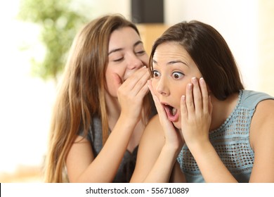 Portrait of a girl telling secrets to her amazed friend sitting on a couch in the living room at home