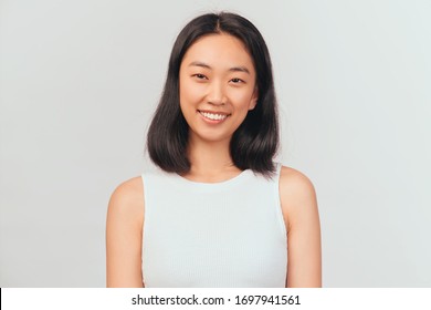 Portrait Of Girl Tilts Head And Smiles Sweetly Showing White Teeth. Beautiful Young Woman Asian Appearance With Black Hair Brown Eyes Stands Isolated White Background In Studio