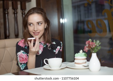 Portrait Of A Girl Using The Voice Recognition Of The Cell Phone And Looking Up Near A Window Of A Restaurant Or A Coffee Shop. People And Technology Concept