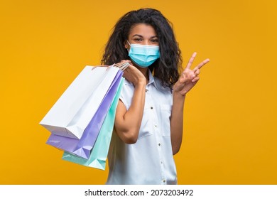 Portrait of a girl wearing protective medical mask on her face, posing with shopper bags and making v-sign gesture, isolated on yellow background. Secure shopping during the quarantine concept.