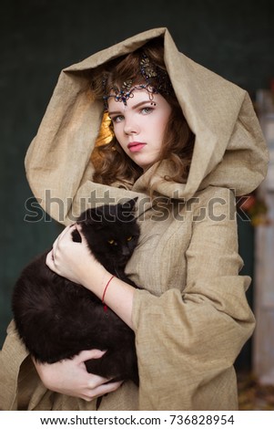 Portrait of a girl, woman, teenage witch in a hood with a black cat on her hands. A pet.