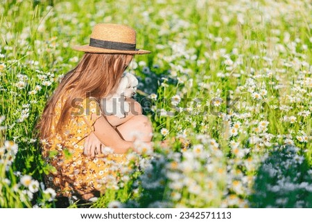 Portrait of girl in a yellow dress and straw hat with cutie small white dog at a chamomile field 