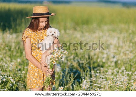 Portrait of girl in a yellow dress and straw hat with cutie small white dog at a chamomile field 