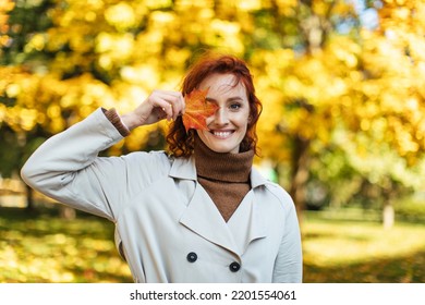 Portrait of glad caucasian millennial red haired female in raincoat puts yellow leaf to her eye, enjoys freedom and warm weather in park in autumn. Ad and offer, walk in city due covid-19 quarantine