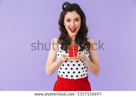 Portrait of glamorous pin-up woman 20s in vintage polka dot dress smiling while holding red present box isolated over violet background